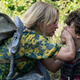 Evelyn Abbott, in a dress covered with yellow flowers and carrying a backpack and a baby sling, clamps a hand over her son's mouth.